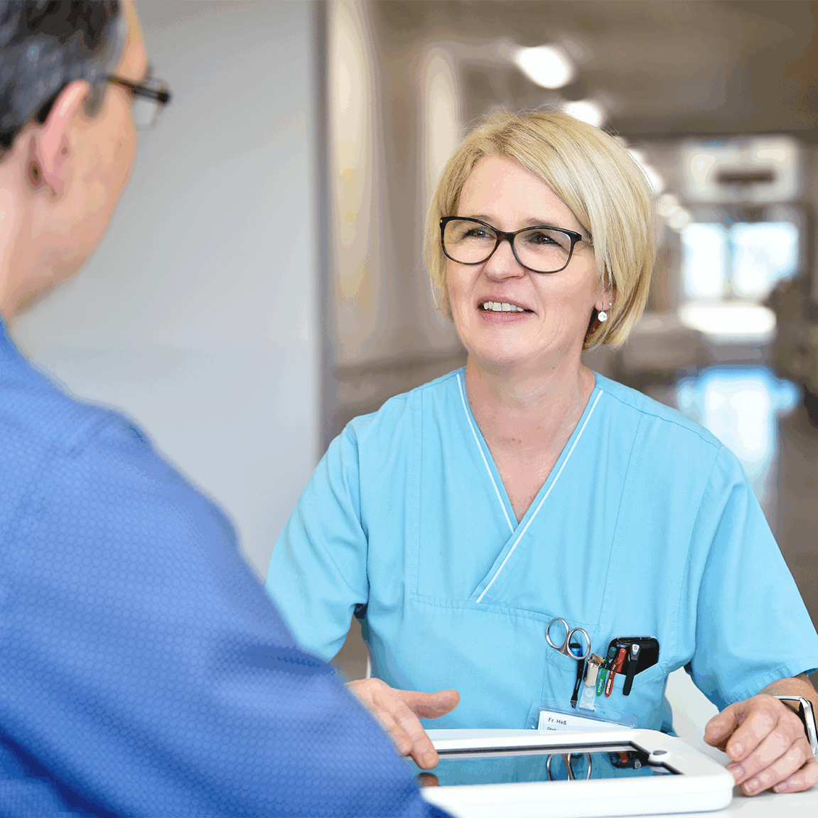 Eine Krankenpflegekraft in blauer Arbeitskleidung spricht mit einem Patienten am Tisch und hält ein Tablet.