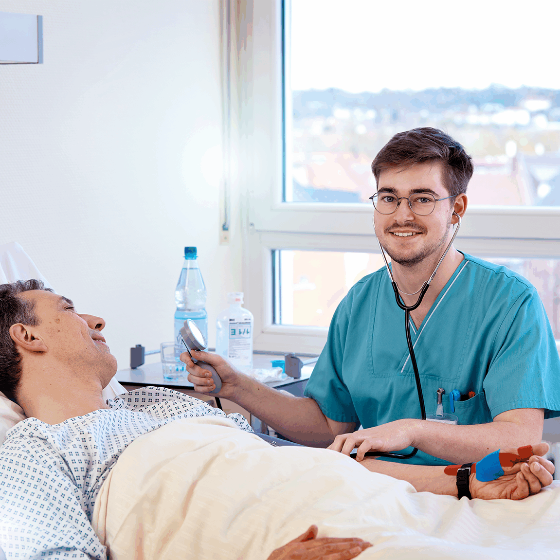 Ein Gesundheits- und Krankenpflegehelfer in blauer Arbeitskleidung untersucht einen lächelnden Patienten im Krankenbett. Im Hintergrund ist ein Fenster mit Blick auf eine Stadtlandschaft zu sehen.