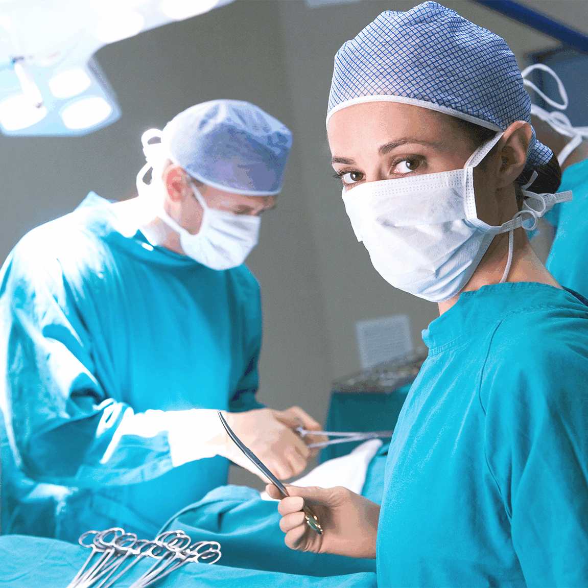Eine Operationsassistentin in grüner OP-Kleidung und Mundschutz blickt in die Kamera, während im Hintergrund weitere Teammitglieder konzentriert an einer Operation arbeiten.